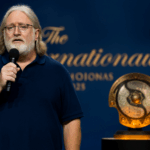 Gabe Newell hablando en The International 2025 en Hamburgo, con el trofeo Aegis of Champions iluminado al fondo.