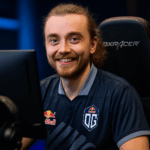 Jugador profesional de OG sonriendo frente al monitor durante un torneo de Dota 2 con luces azules y naranjas